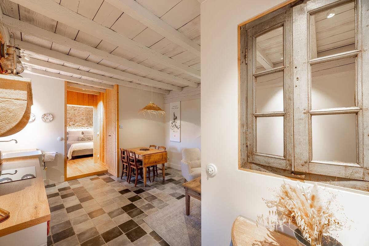 Une pièce avec un sol à carreaux, des poutres en bois au plafond, une table à manger en bois avec des chaises, un lit et une fenêtre avec un cadre en bois.