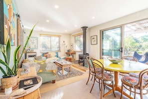 A cozy living room with a sofa, coffee table, and dining table. There is a fireplace and a large window.