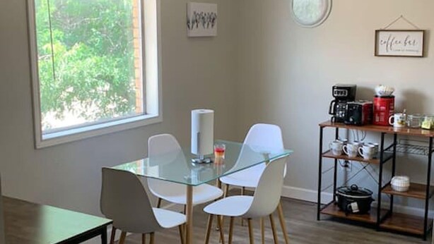 A dining area with a glass table and white chairs, a coffee station with a coffee maker and mugs, and a window with a view of greenery.