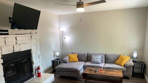 A living room with a fireplace, a grey sofa with yellow cushions, a wooden coffee table, and a flat-screen TV mounted on the wall.