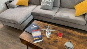 A grey sectional sofa with two yellow pillows, a wooden coffee table with a book titled 'FAR AIM 2017', a small airplane model, and a candle.