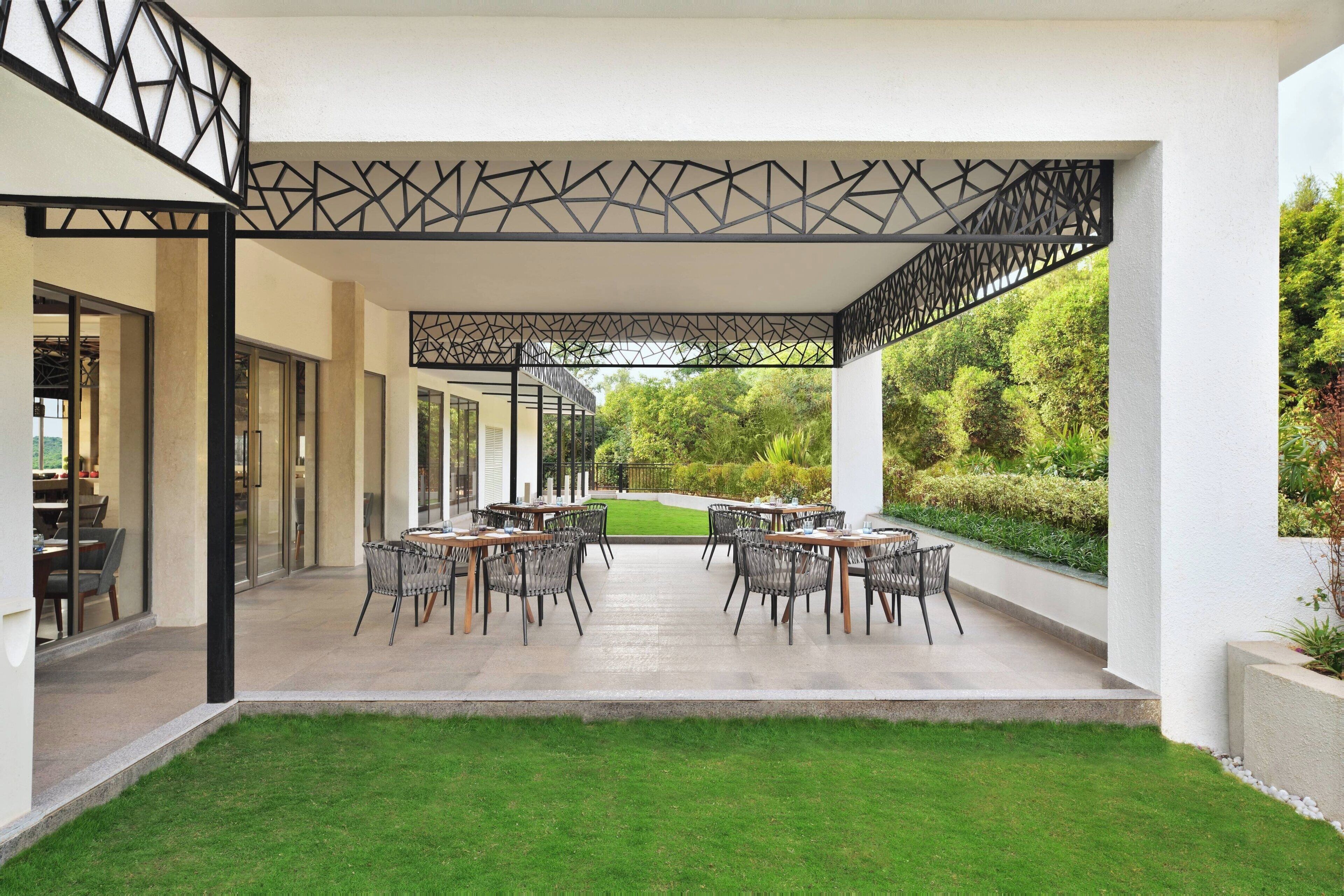 Un patio al aire libre con un diseño geométrico en el techo, césped y una zona de comedor con mesas y sillas.