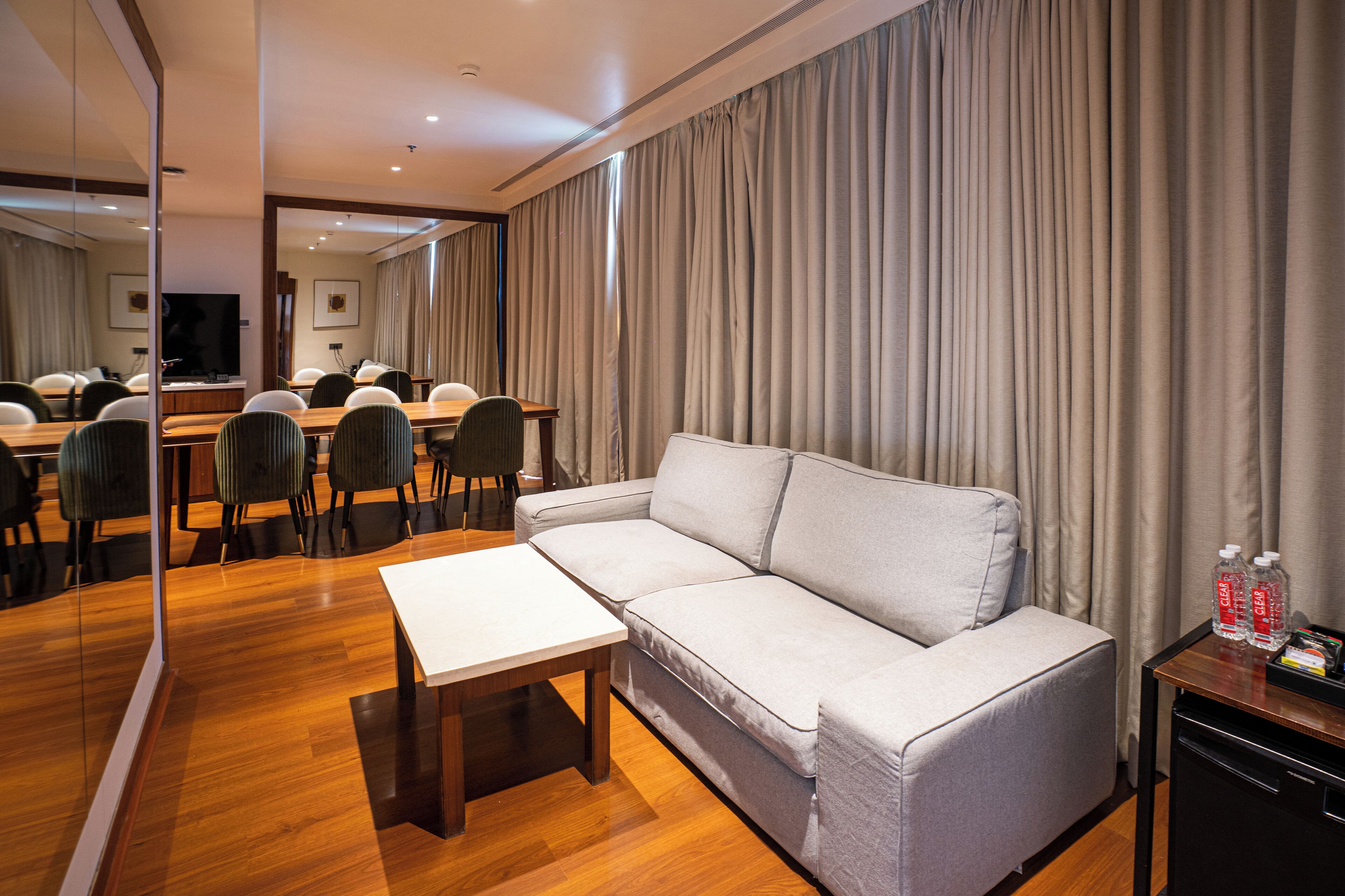 Une chambre d’hôtel moderne équipée d’un canapé, d’une petite table, d’un bureau avec des chaises, d’une télévision et d’un miroir.