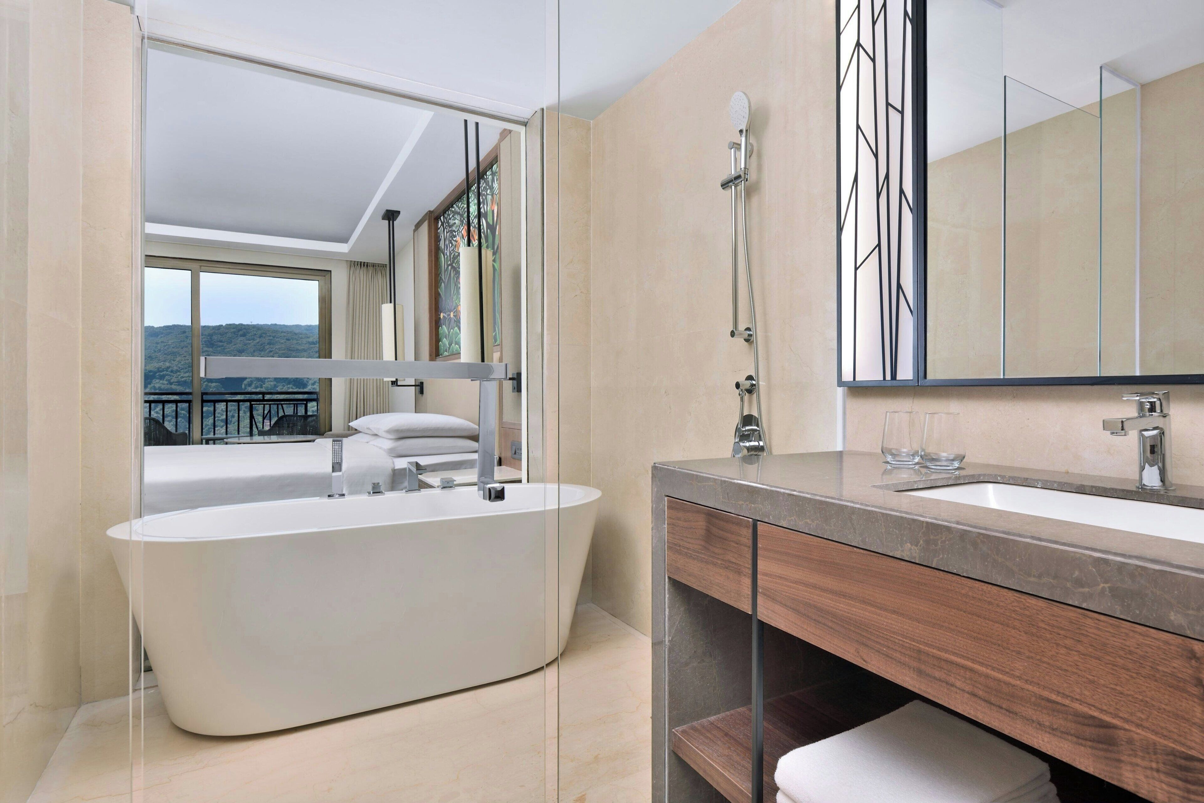 A modern bathroom with a freestanding bathtub, a large mirror, and a view of a balcony with a scenic landscape.