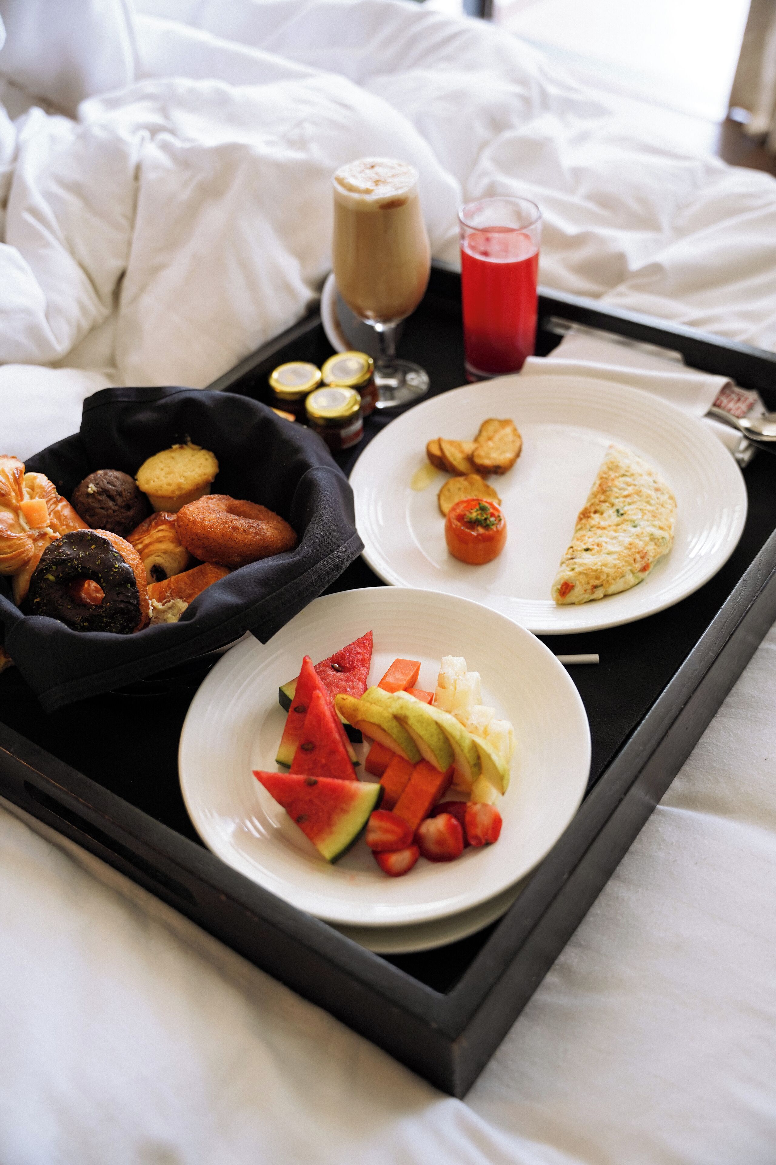 A breakfast tray with various foods including toast, a slice of omelette, a glass of red juice, and a bowl of assorted pastries.