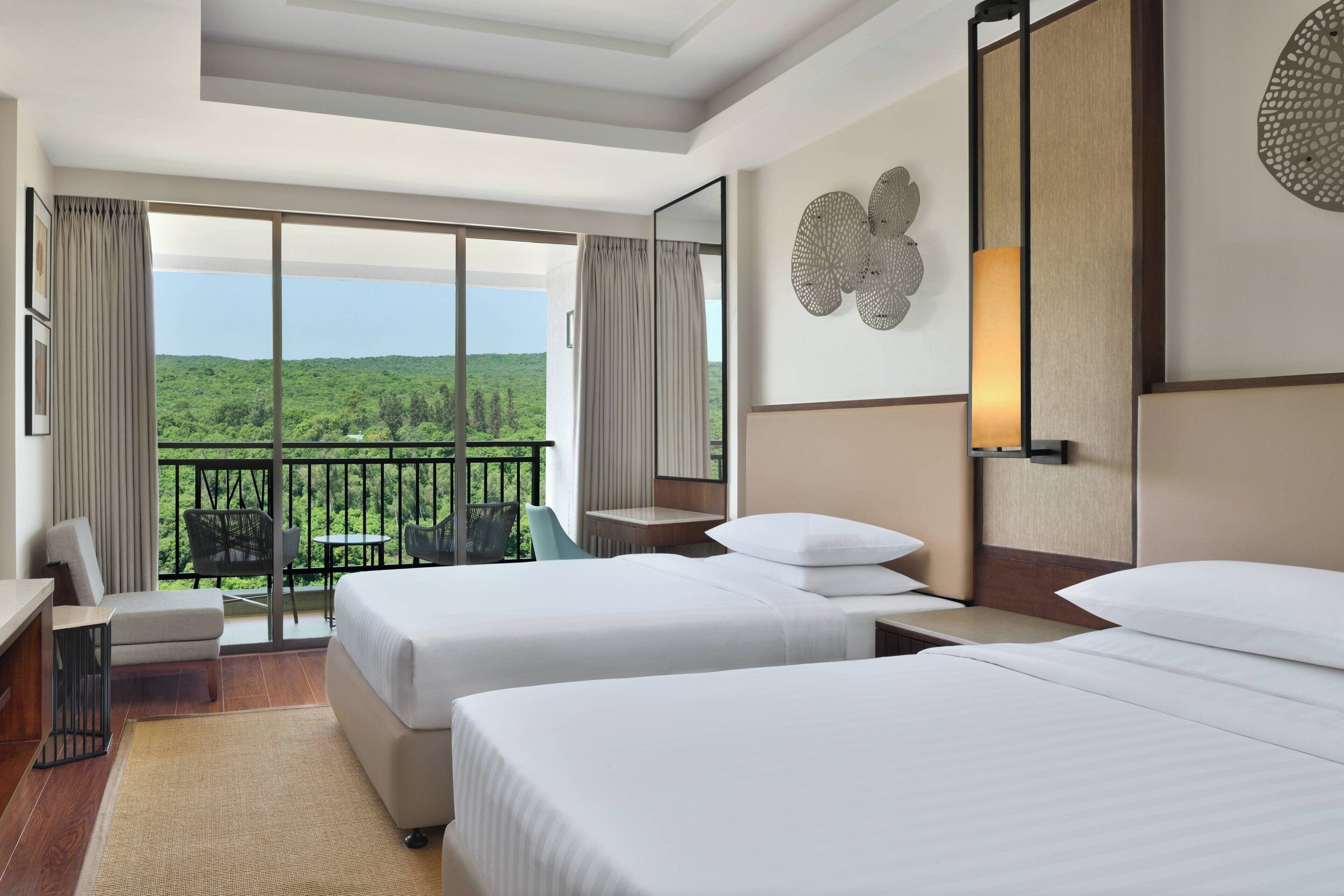 A hotel room with two beds, a desk, a chair, and a balcony with a view of greenery.