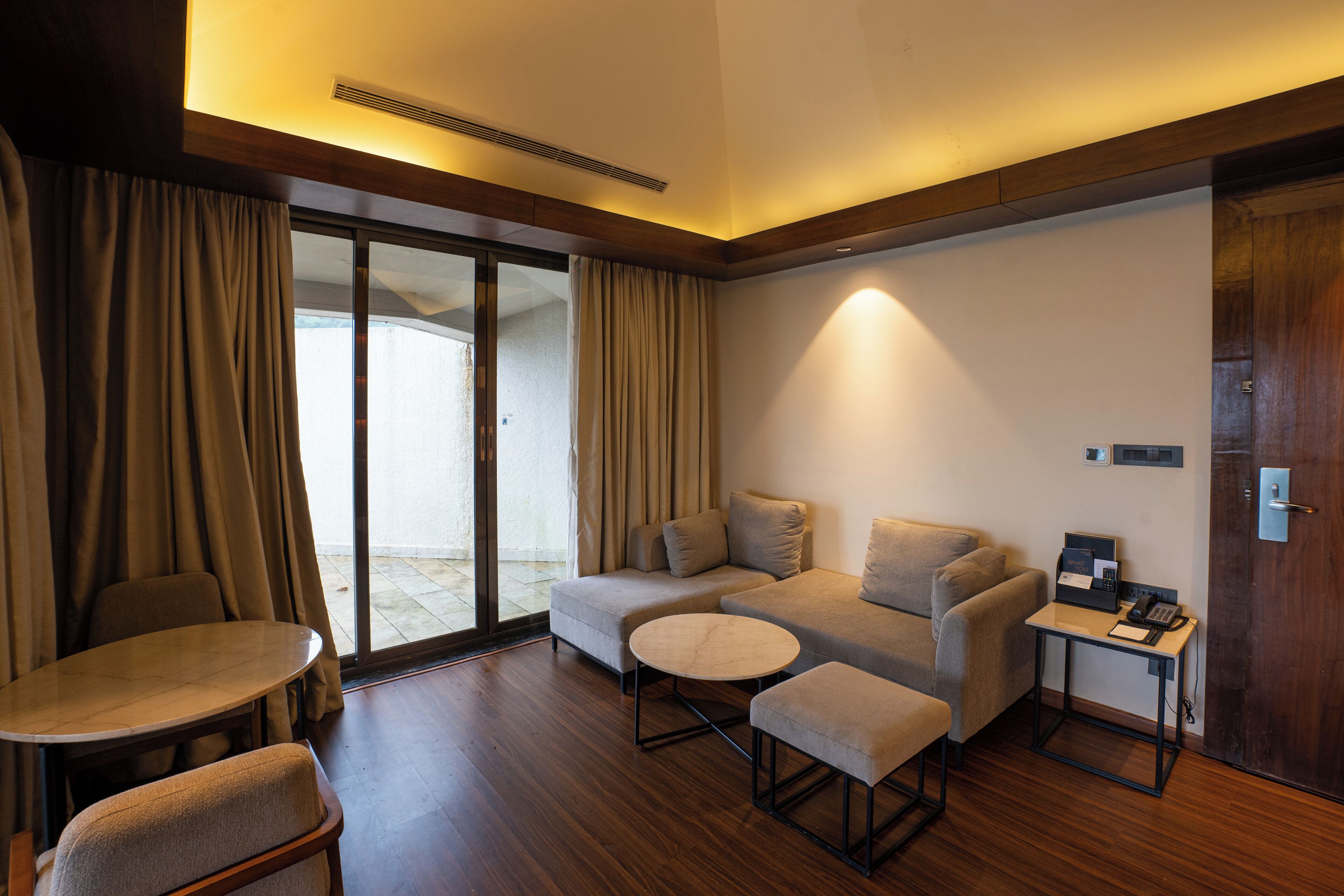 A modern hotel room with a sofa, a round table, and a small desk with a phone and a book.