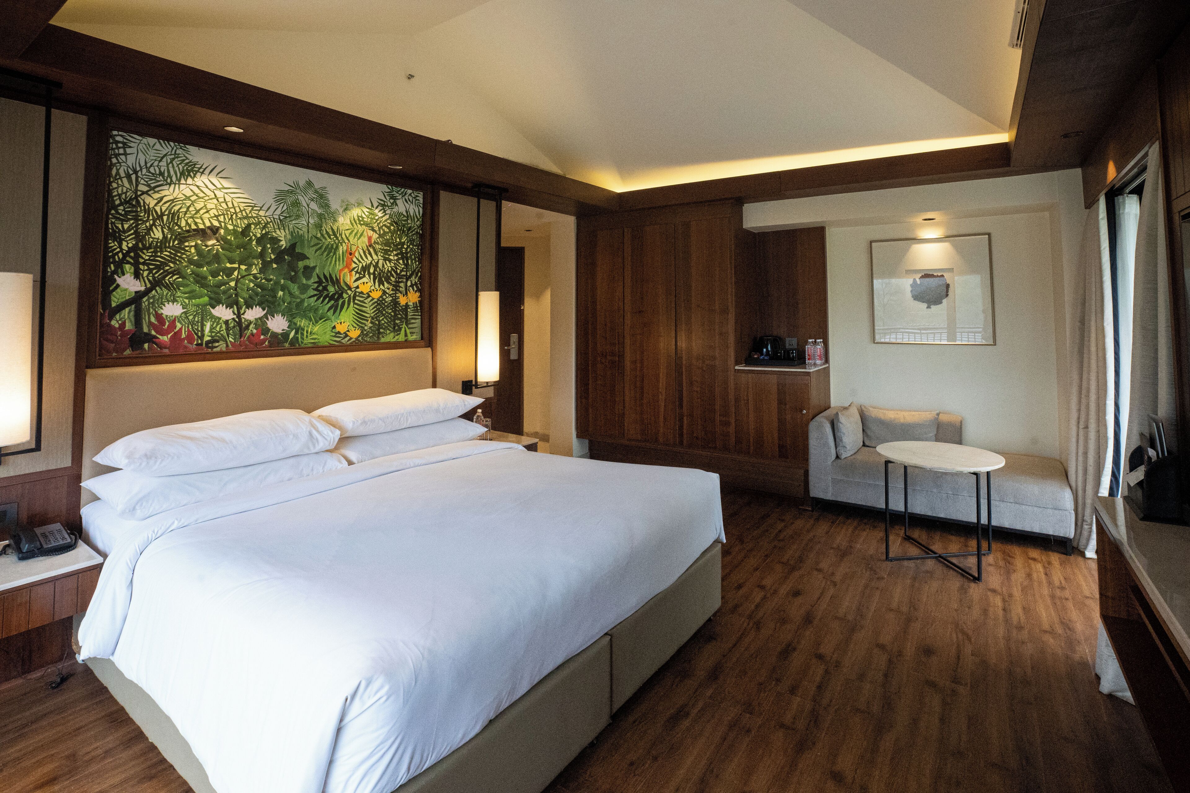 A hotel room with a large bed, a small table, a sofa, a painting, and a kitchenette.