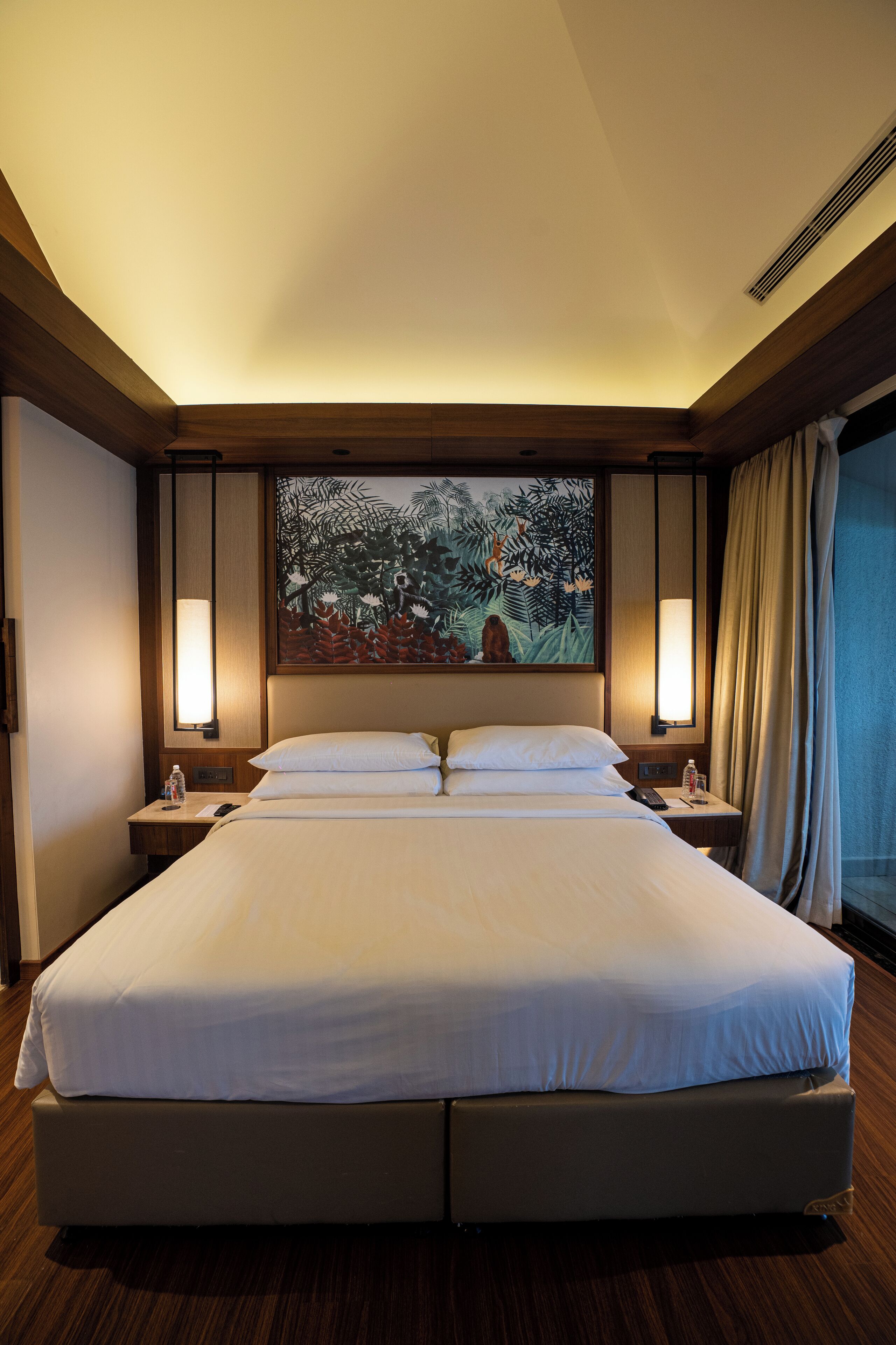 A large bed with white linens, two bedside tables with lamps, a painting above the bed, and a window with curtains.