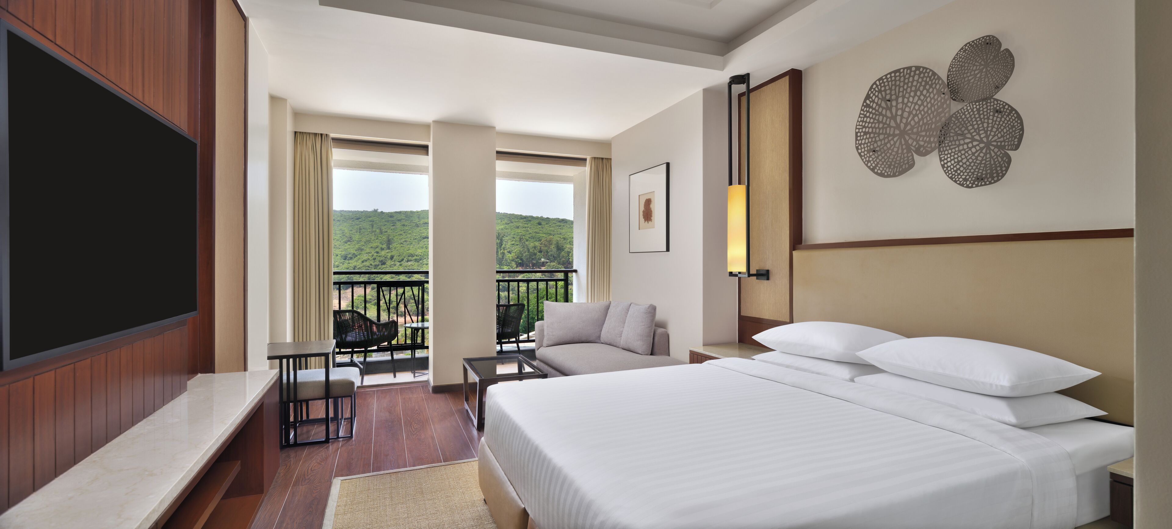 A hotel room with a large bed, a TV, a balcony with outdoor seating, and a view of greenery.