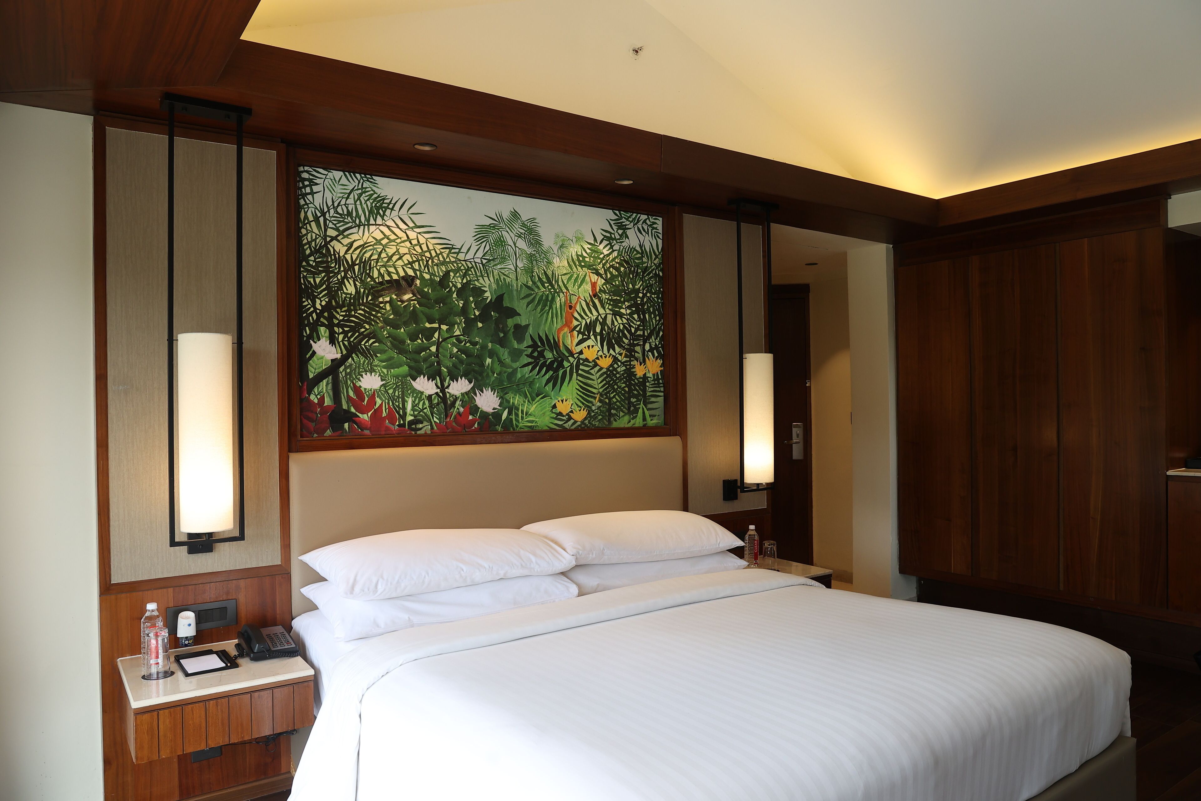 A hotel room with a large bed, wooden headboard, and a wall mural of tropical foliage. There are two bedside lamps and a nightstand with a phone and a bottle.