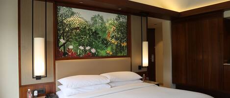 A hotel room with a large bed, wooden headboard, and a wall mural of tropical foliage. There are two bedside lamps and a nightstand with a phone and a bottle.