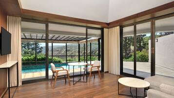 Una habitación moderna con piso de madera, una mesa redonda con sillas, un sofá, un televisor en la pared y ventanas amplias con vista a una piscina y follaje.