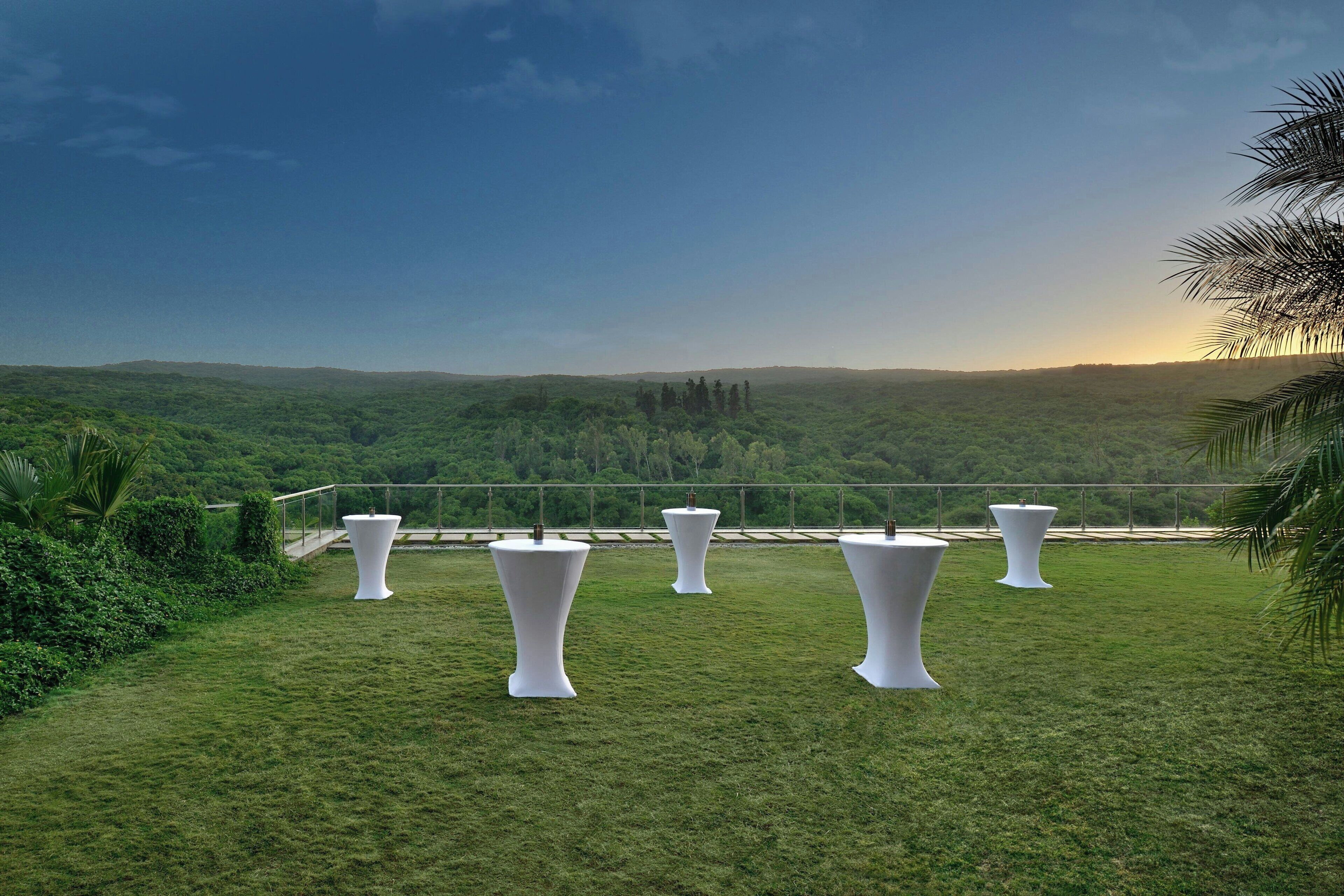 A grassy outdoor area with white tables and chairs, surrounded by greenery and a scenic view of hills.