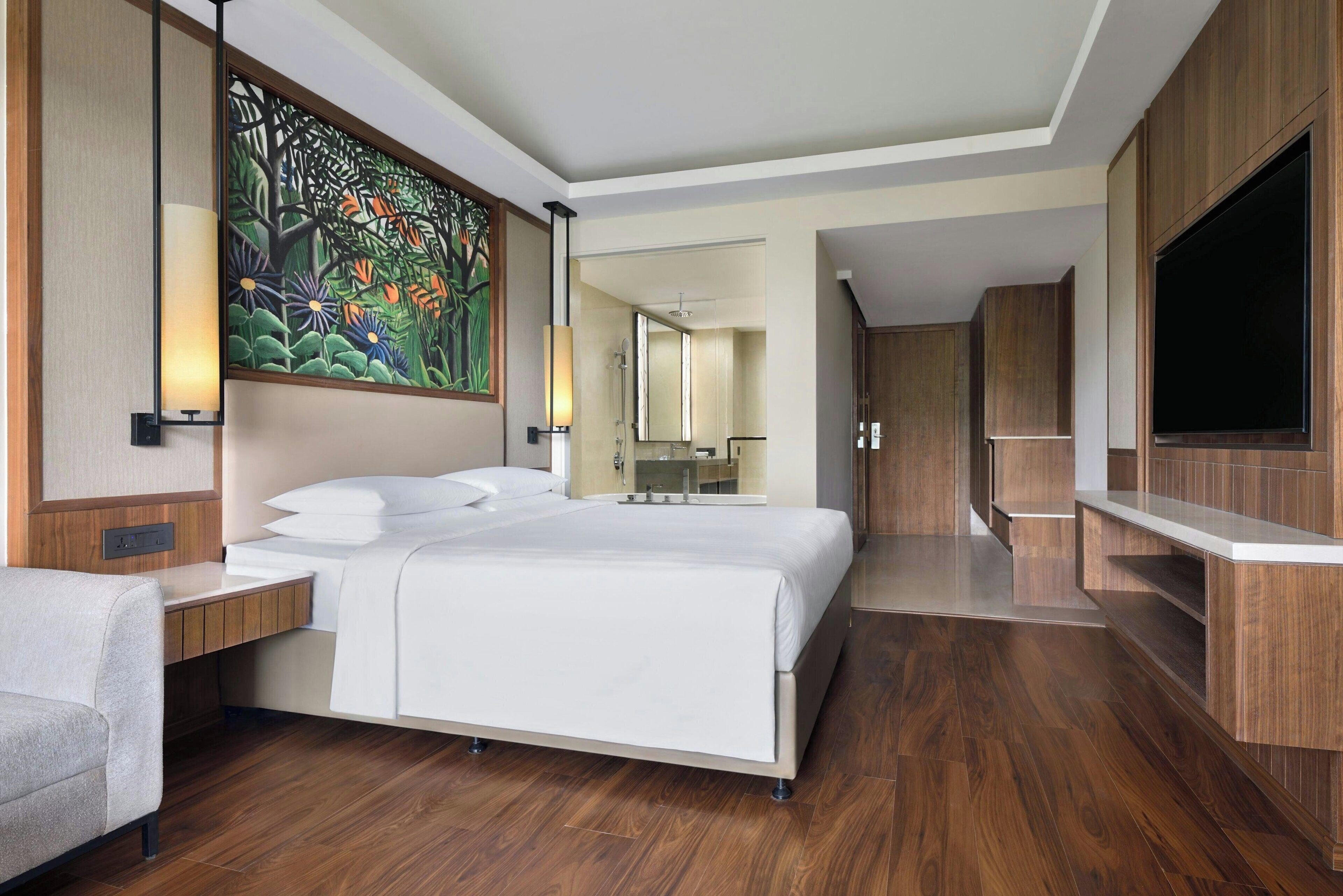 Quarto de hotel moderno com uma cama grande, piso de madeira, televisão e banheiro visível através de uma porta aberta.