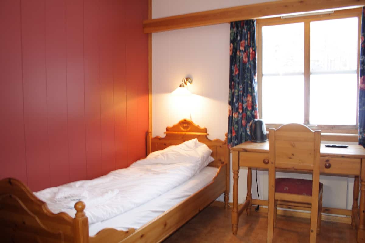 Una cama individual con estructura de madera, un escritorio con silla y una ventana con cortinas.