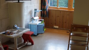 A room with a play area including a table with toys, a small blue playhouse, and a red stool. There is a window with curtains and a wooden floor. - Dovreskogen Gjestegård  (Dovre)