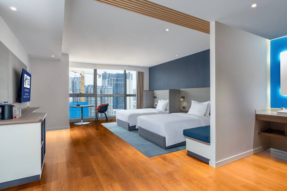 A modern hotel room with two beds, a desk, a bathroom with a mirror, and a city view.