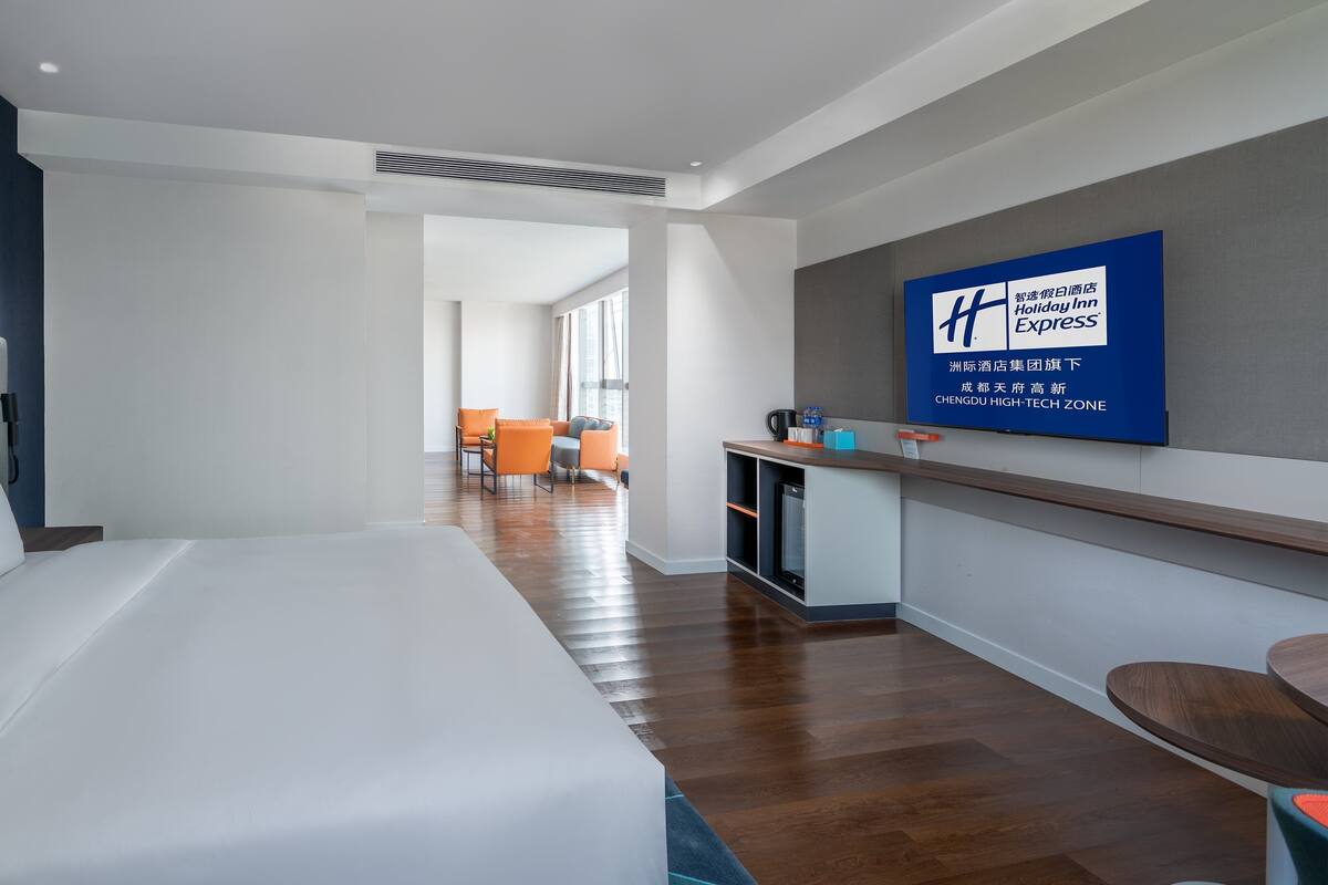 A modern hotel room with a large bed, a desk, and a seating area with orange chairs.