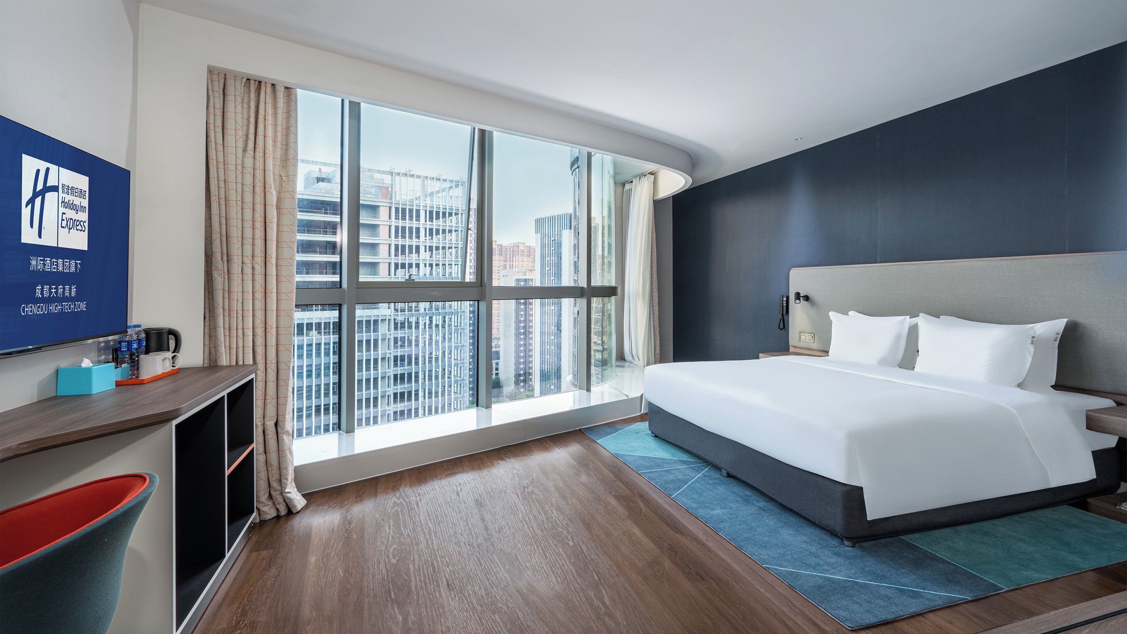 A hotel room with a large window, a bed, a desk, and a flat-screen TV.