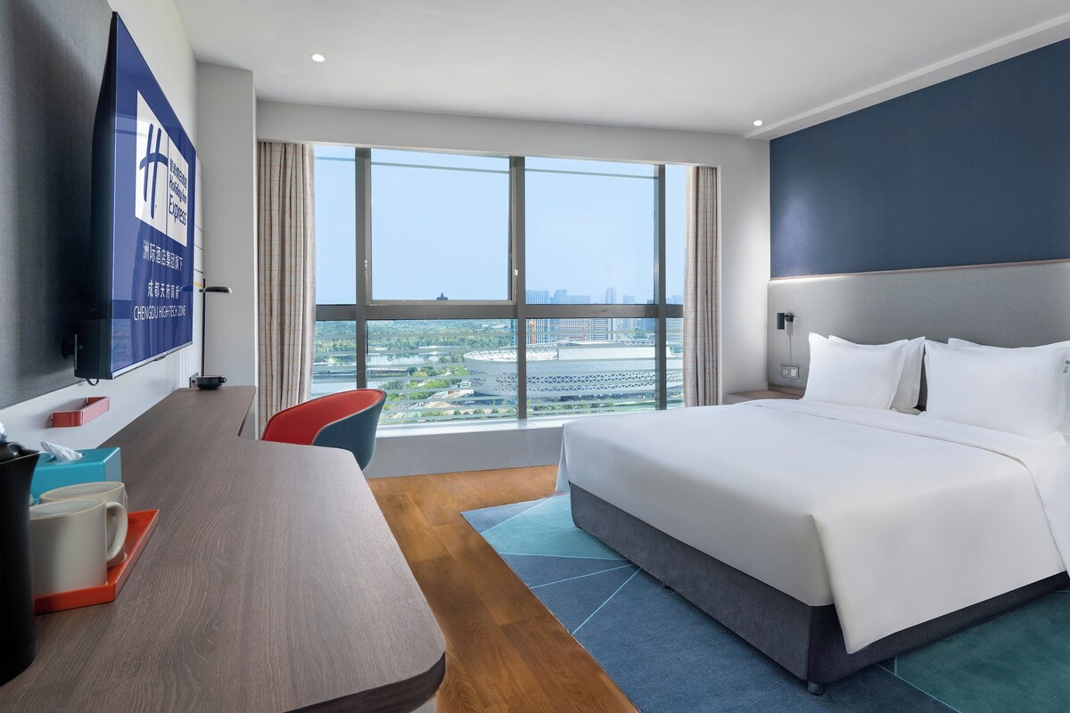 Ett hotellrum med en stor säng, ett skrivbord med en stol, en TV och utsikt över stadsbilden genom stora fönster.