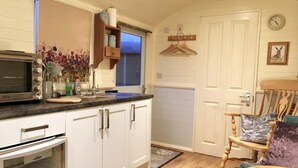 A compact kitchen with white cabinets, a microwave, and a small oven. There's a wooden chair with a cushion and a small rug on the floor. - Meadowsweet - luxury Shepherd's Hut, amazing views (Herstmonceux)