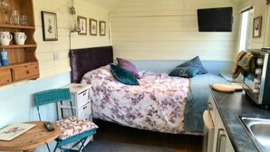 A compact, well-lit room with a bed, a small table with chairs, a kitchenette, and a shelf with glassware. - Meadowsweet - luxury Shepherd's Hut, amazing views (Herstmonceux)