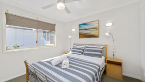 Un dormitorio con cama, ventana con persianas, ventilador de techo, lámpara de noche, mesita de noche y un cuadro en la pared.