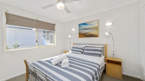 A bedroom with a bed, a window with blinds, a ceiling fan, a bedside lamp, a nightstand, and a painting on the wall. - Bimbimbi 1, 11 Willow Street (Crescent Head)