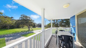 Un espacio al aire libre cubierto con una mesa, sillas, barandilla blanca y vista a un césped verde y árboles.
