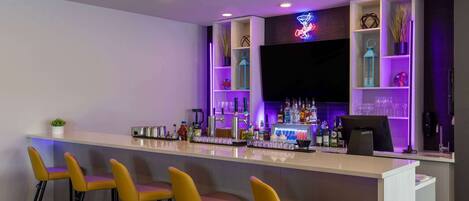 A modern bar area with a white countertop, four yellow bar stools, and shelves with bottles and glasses.