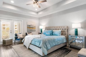 A bedroom with a large bed, a ceiling fan, a nightstand with a lamp, and a window with blinds.