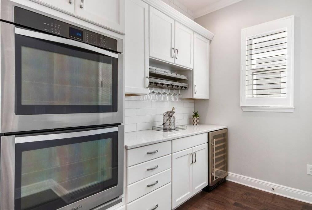 Una cocina con dos hornos de acero inoxidable, armarios blancos, una encimera con un recipiente de vidrio y una ventana con persianas.