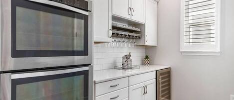 Una cocina con dos hornos de acero inoxidable, armarios blancos, una encimera con un recipiente de vidrio y una ventana con persianas.