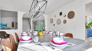 Un'elegante sala da pranzo con un lungo tavolo apparecchiato per sei persone, circondato da sedie. Si notano diverse pendole a parete, un lampadario sopra il tavolo e una pianta.