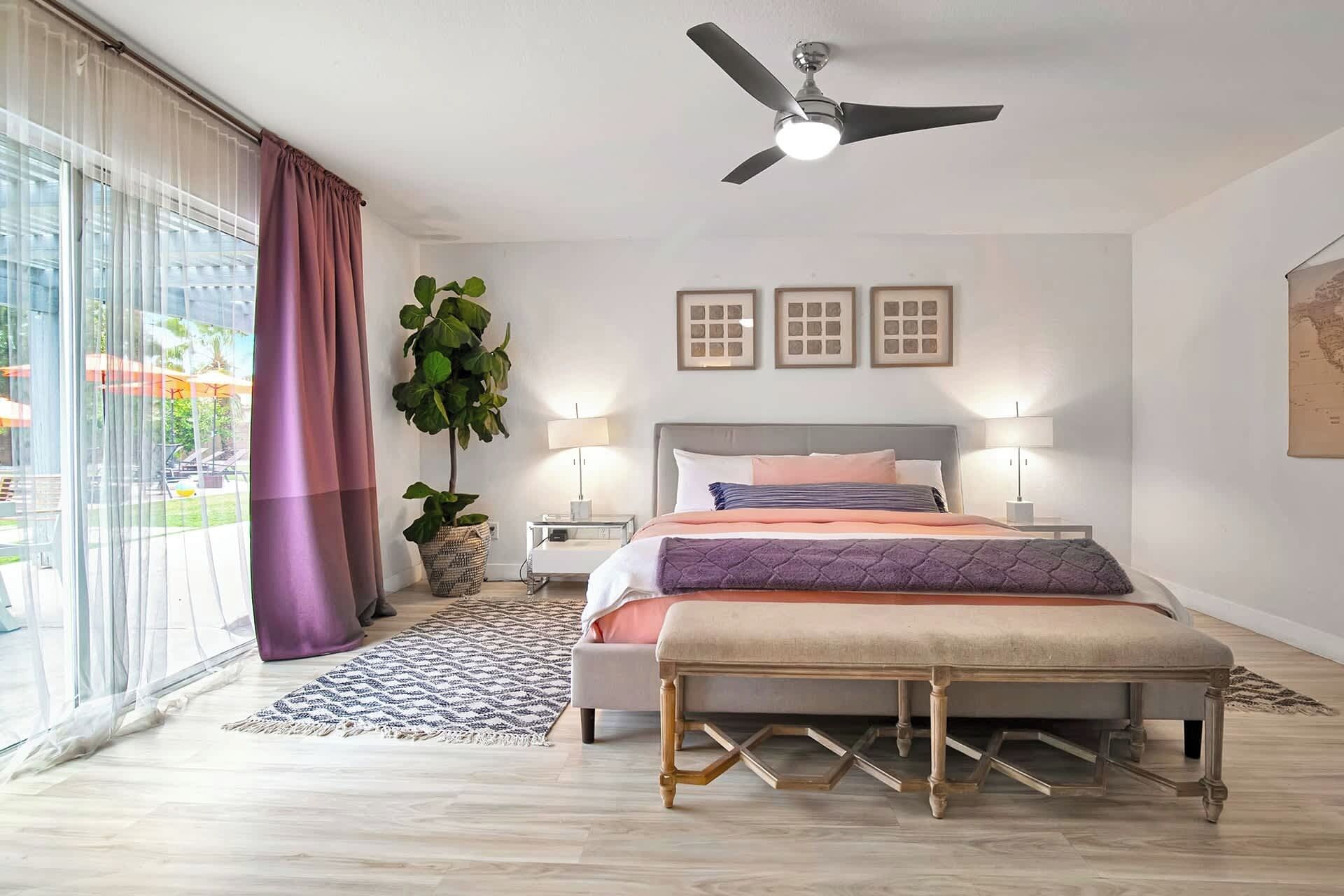 A bedroom with a bed, bedside tables, a ceiling fan, a large window with curtains, and a potted plant.