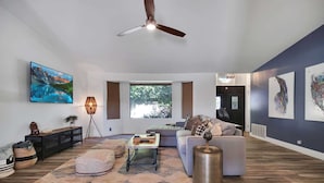A spacious living room with a large window, a ceiling fan, a flat-screen TV, and a sofa.