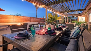 An outdoor patio area with wooden pergola, dining tables, and chairs, surrounded by plants and a wooden fence.