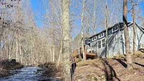 A rustic cabin with a wooden deck overlooking a flowing river. - Waterfall Cottage | Hot Tub, Game Room, Fireplace (Mentone)