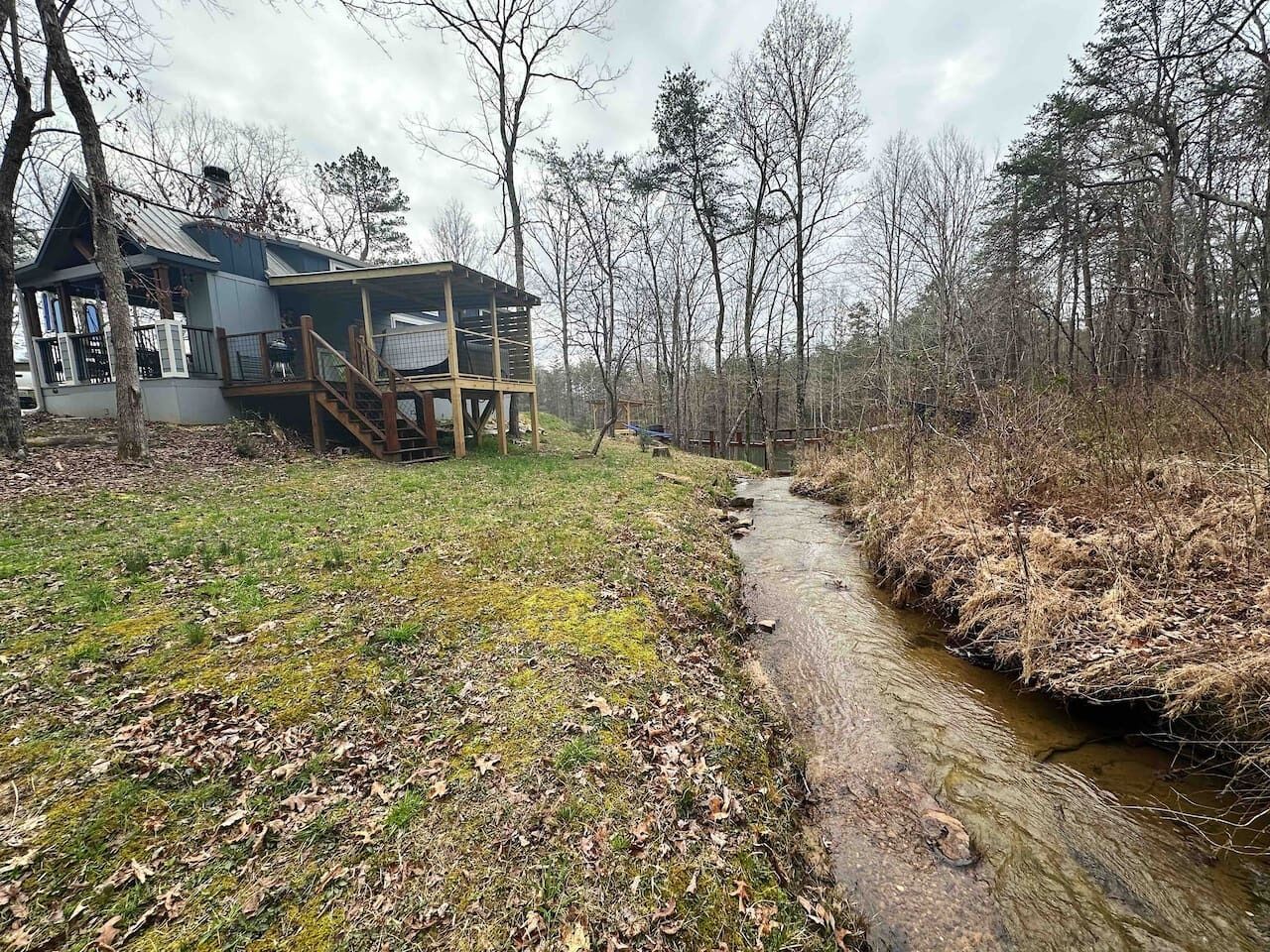 A rustic cabin with a wooden deck and a small stream running beside it.