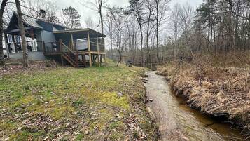 A rustic cabin with a wooden deck and a small stream running beside it.