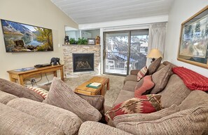 A cozy living room with a fireplace, a large window with a view of a deck, and a painting of a mountain landscape.