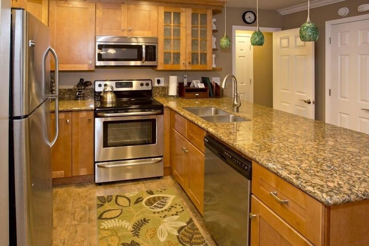 A kitchen with wooden cabinets, stainless steel appliances, a granite countertop, and a patterned rug.