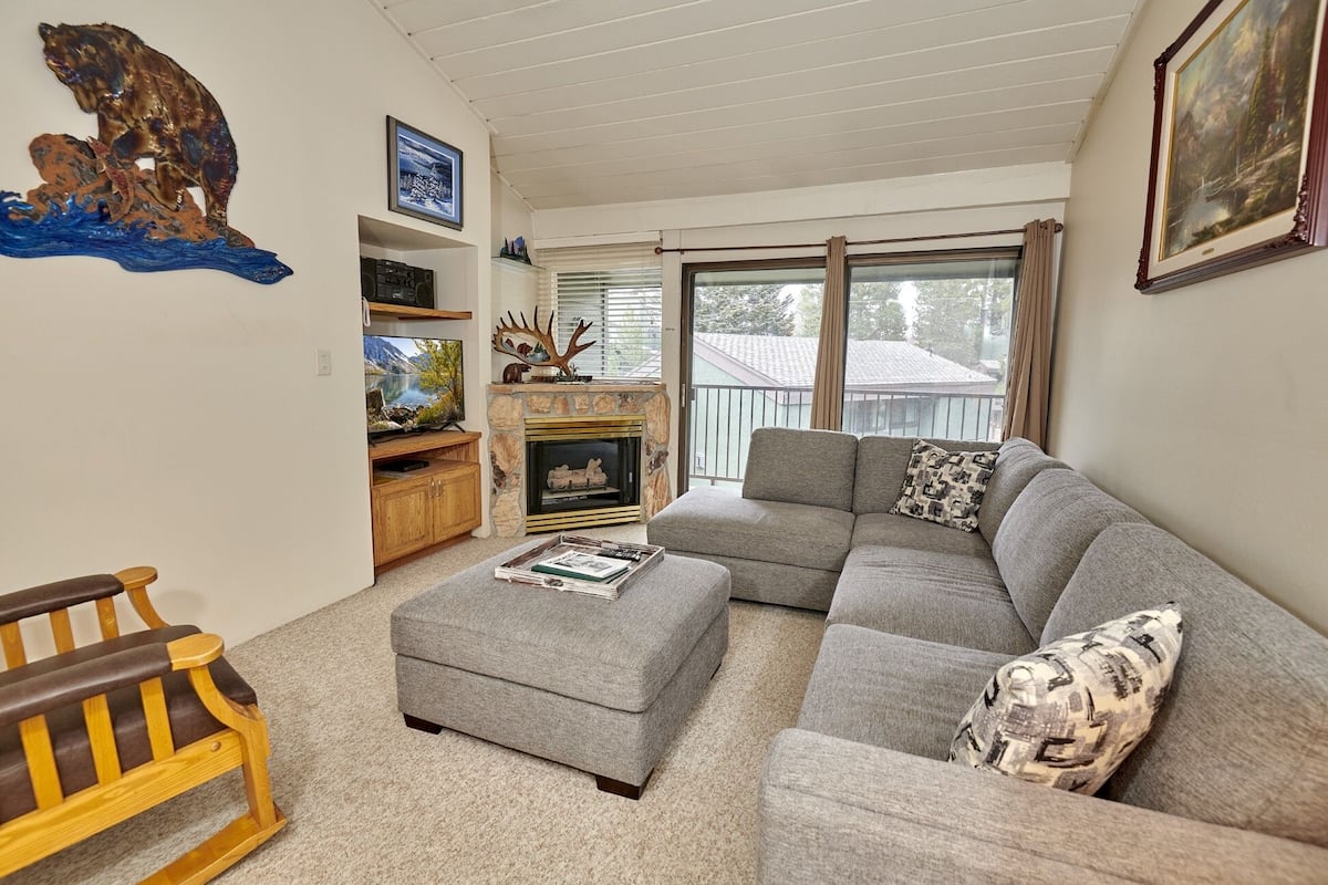 A living room with a stone fireplace, a grey sectional sofa, a wooden bench, and a television on a wooden stand.