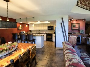 A modern kitchen with white cabinets, a central island, and a dining area with a wooden table and chairs. - Epic Mountain Views! End Unit! 2 Miles North of Purgatory! Heated Pool ! (durango)