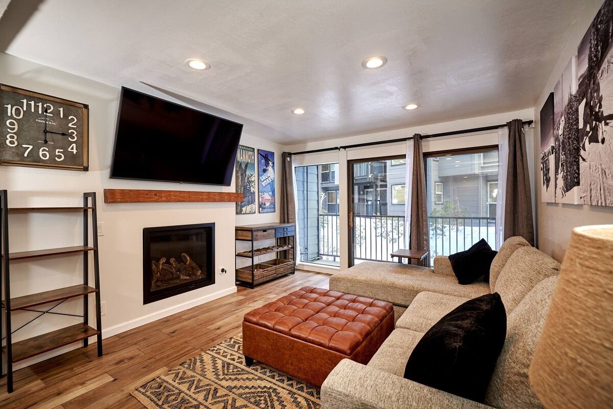 A living room with a sofa, ottoman, TV, fireplace, and a large window with a view of buildings.