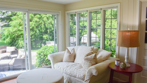 A cozy living room with a white sofa, a round ottoman, and a small table with a lamp. Large windows provide a view of a garden and outdoor furniture. - Lovely three bedroom home located in desirable West Dennis neighborhood! (West Dennis)