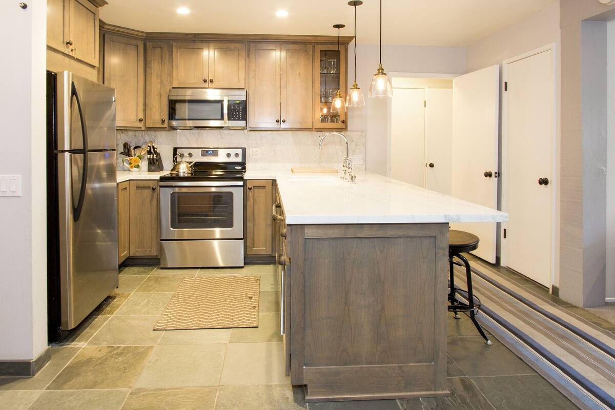 A modern kitchen with wooden cabinets, stainless steel appliances, a marble countertop, and pendant lighting.