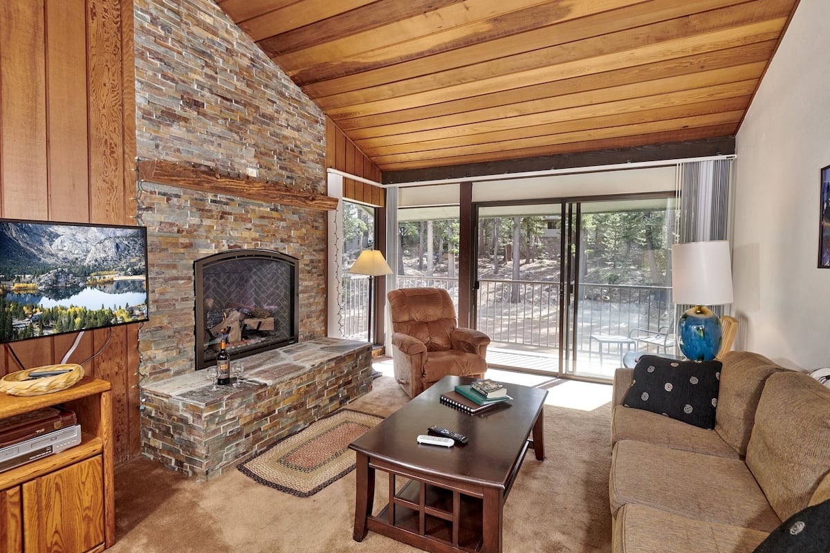 A cozy living room with a stone fireplace, wooden ceiling, and large windows offering a view of trees.