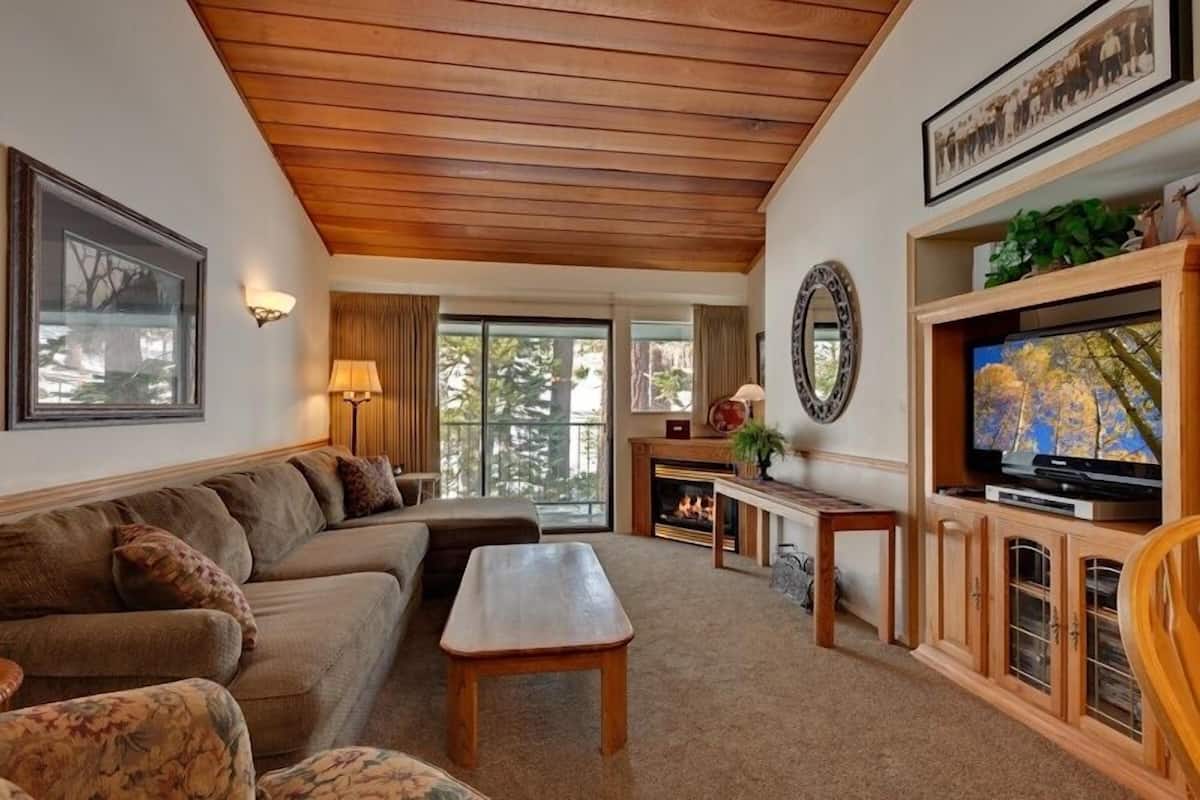 A cozy living room with a wooden ceiling, a fireplace, a television, and a large window.